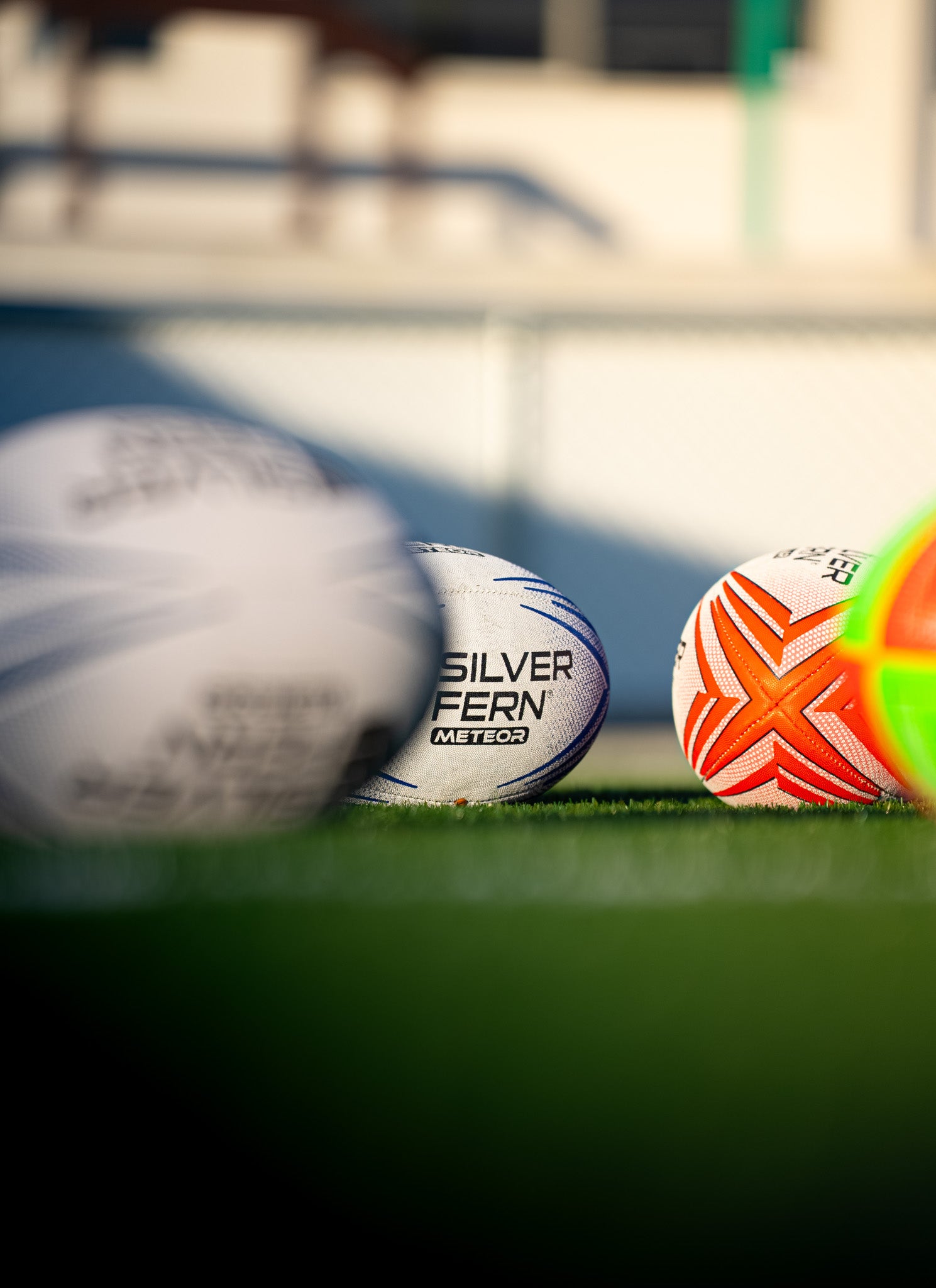 Three rugby balls on green grass in a sports field, with one ball in sharp focus showing the text 'SILVER FERN METEOR' and another ball partially visible in the foreground that is heavily blurred.